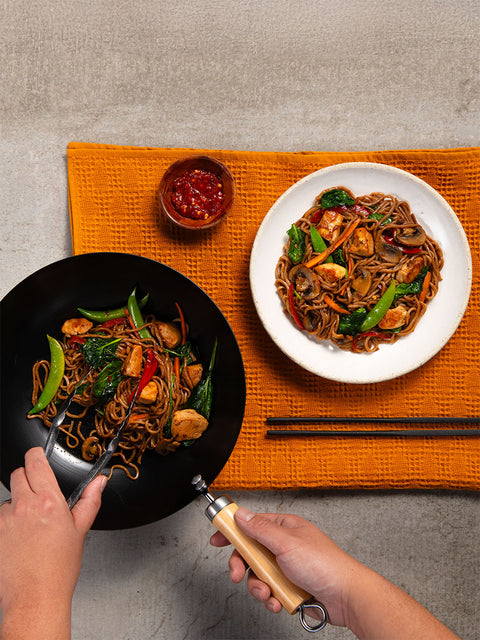 Person cooking and plating easy chicken lo mein on an orange placemat, with a hand using chopsticks with nuPasta noodles.