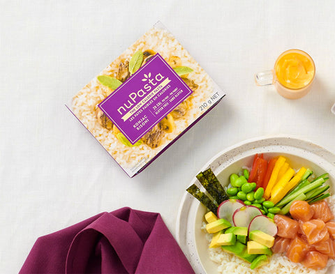 Box of nuPasta Risoni with a bowl of salad and a glass of juice on a white surface