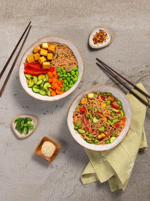 Two bowls of spring noodle salad with vegetables on a gray surface with chopsticks and small dishes made with nuPasta noodles.